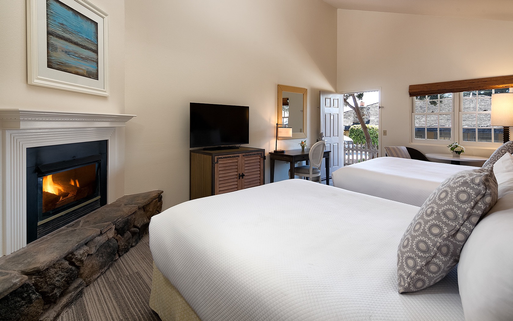 A hotel room with a fireplace, two beds, a TV, a desk, and a window